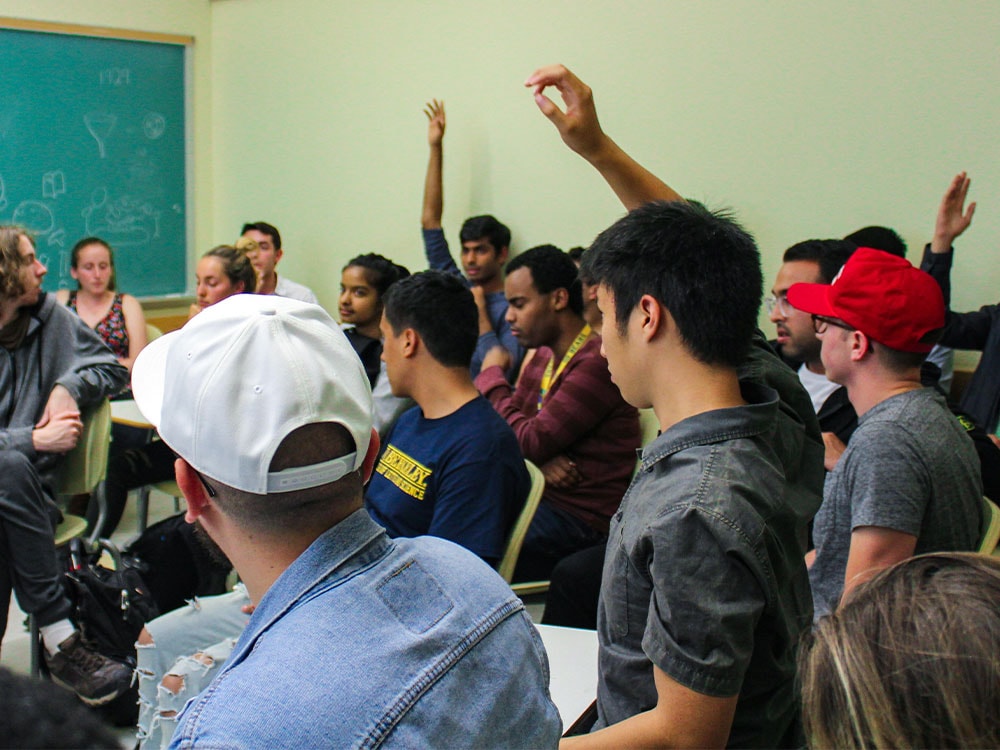 A diverse group of students attending a Bridge USA discussion in a classroom. Many of the students have their hands raised.